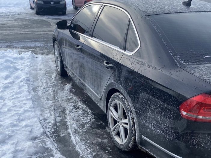 Black sedan parked on a snowy, icy street with snow piled along the curb; frost and water droplets on the car’s windows and body.