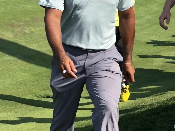 Golfer in a light blue polo and gray pants stepping on a grassy golf course, wearing a white cap and white Nike shoes.
